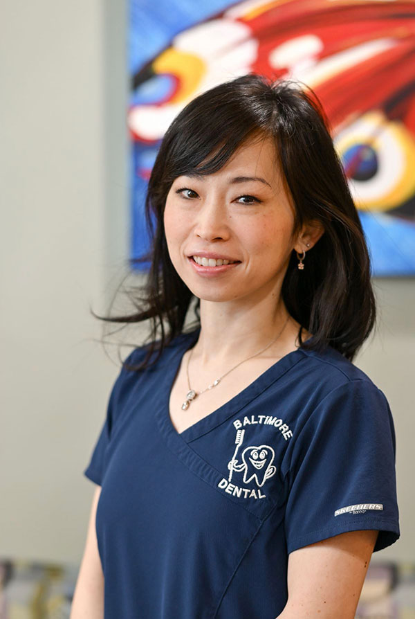 The image shows a woman in a blue scrub uniform, smiling and posing for the photograph. She is wearing a name tag with the text  Albrighton Dental.