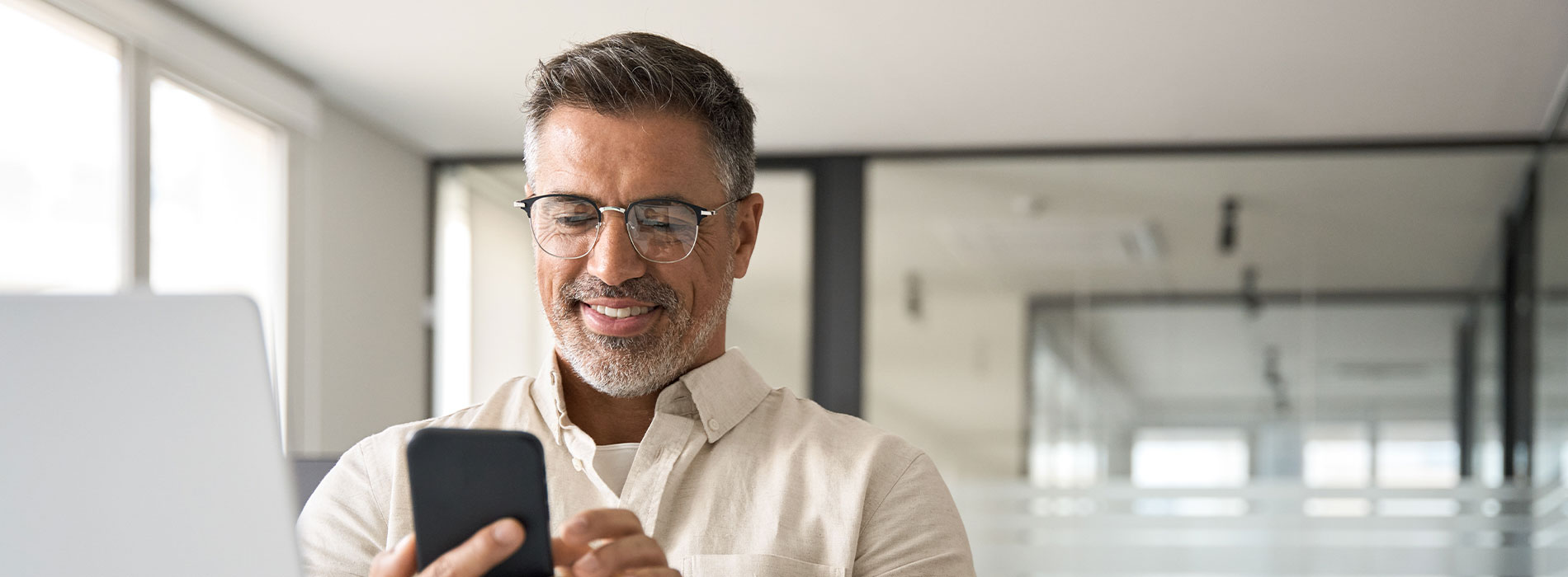 A man in a professional setting, holding a phone and looking at it with a smile.