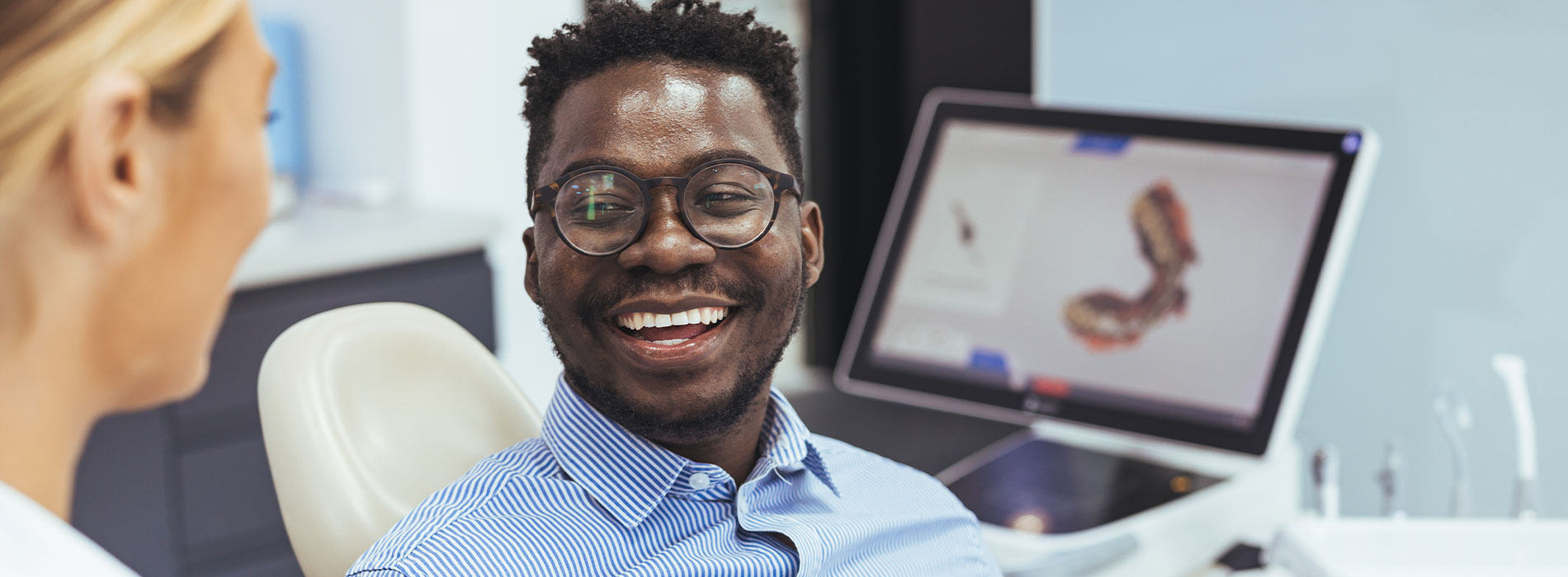 A man is sitting in front of a computer screen, smiling and looking at the camera.