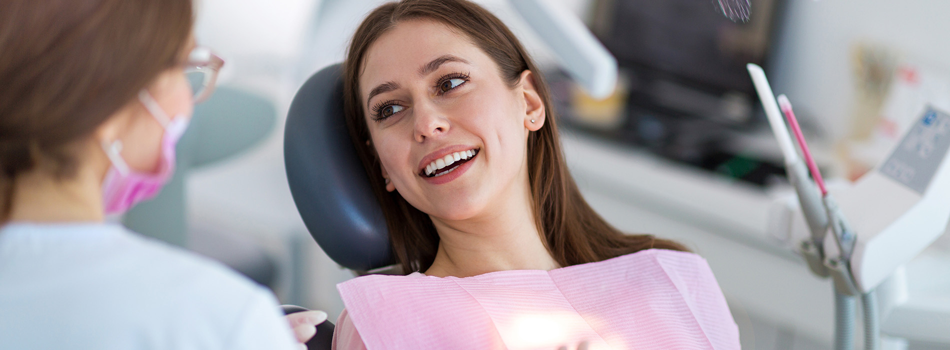 The image shows a woman in a dental office setting, smiling and sitting in a dentist s chair while being attended to by a female dental professional.