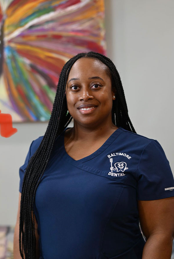The image is a photograph of a woman standing indoors. She has dark hair, wears glasses and a blue scrub top with the word  Baltimore  visible on it, suggesting she may be a healthcare professional.