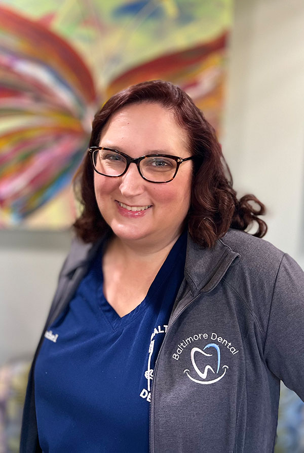 The image is a photograph of a woman standing in front of an abstract painting. She is wearing glasses, a dark top, and has short hair. Her attire suggests she may be a professional, possibly in the medical field, indicated by her name tag which reads  Dental Office.