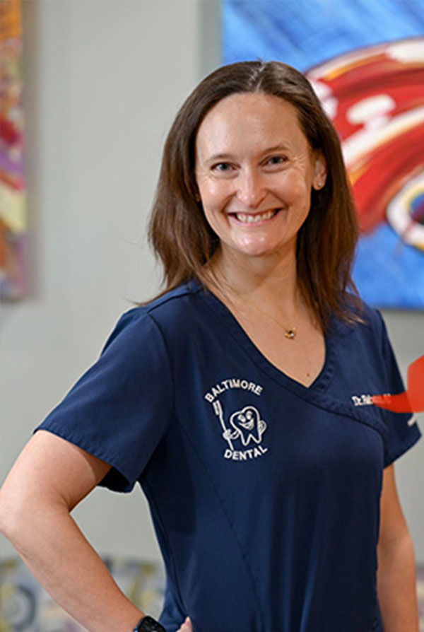 The image is a portrait of a woman wearing a blue scrub top with a logo on the left chest area, standing in front of a colorful wall and smiling at the camera.