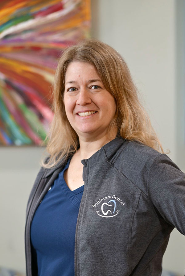 A woman in a blue shirt and a jacket with a logo, standing in front of a painting.