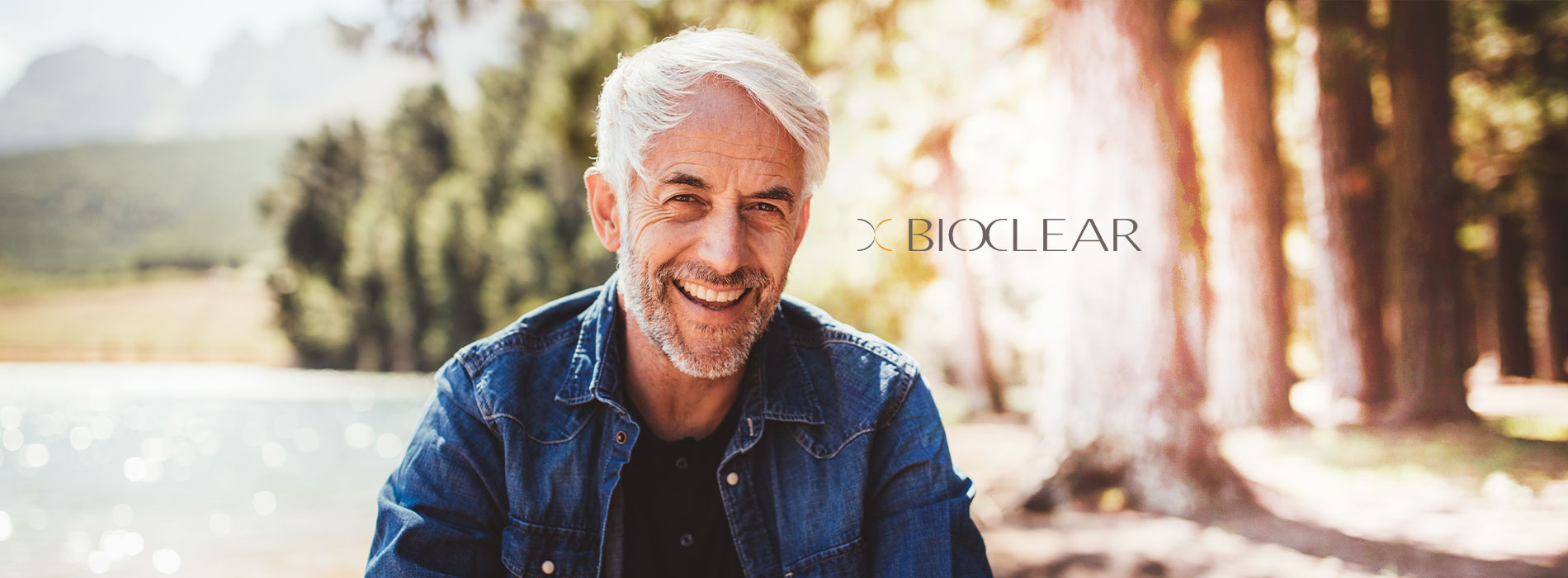 The image shows a man with gray hair, wearing a denim jacket, sitting outdoors in front of trees. He is smiling and appears to be in a relaxed posture.