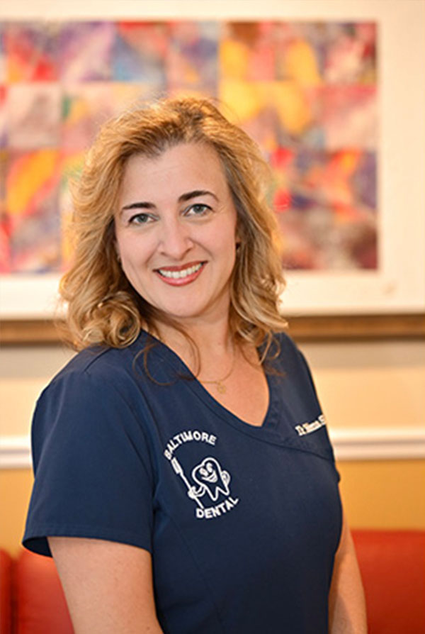 The image is a photograph of a woman posing for the camera. She appears to be in a professional setting, possibly an office or a reception area, as indicated by the presence of a framed picture on the wall behind her and a piece of artwork with a colorful abstract design. The woman has short brown hair, is wearing a blue shirt with a name tag that reads  Dental,  suggesting she may be a dental professional, and has a smile on her face.