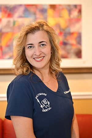 Woman in blue scrubs standing in front of a framed artwork, smiling at the camera.