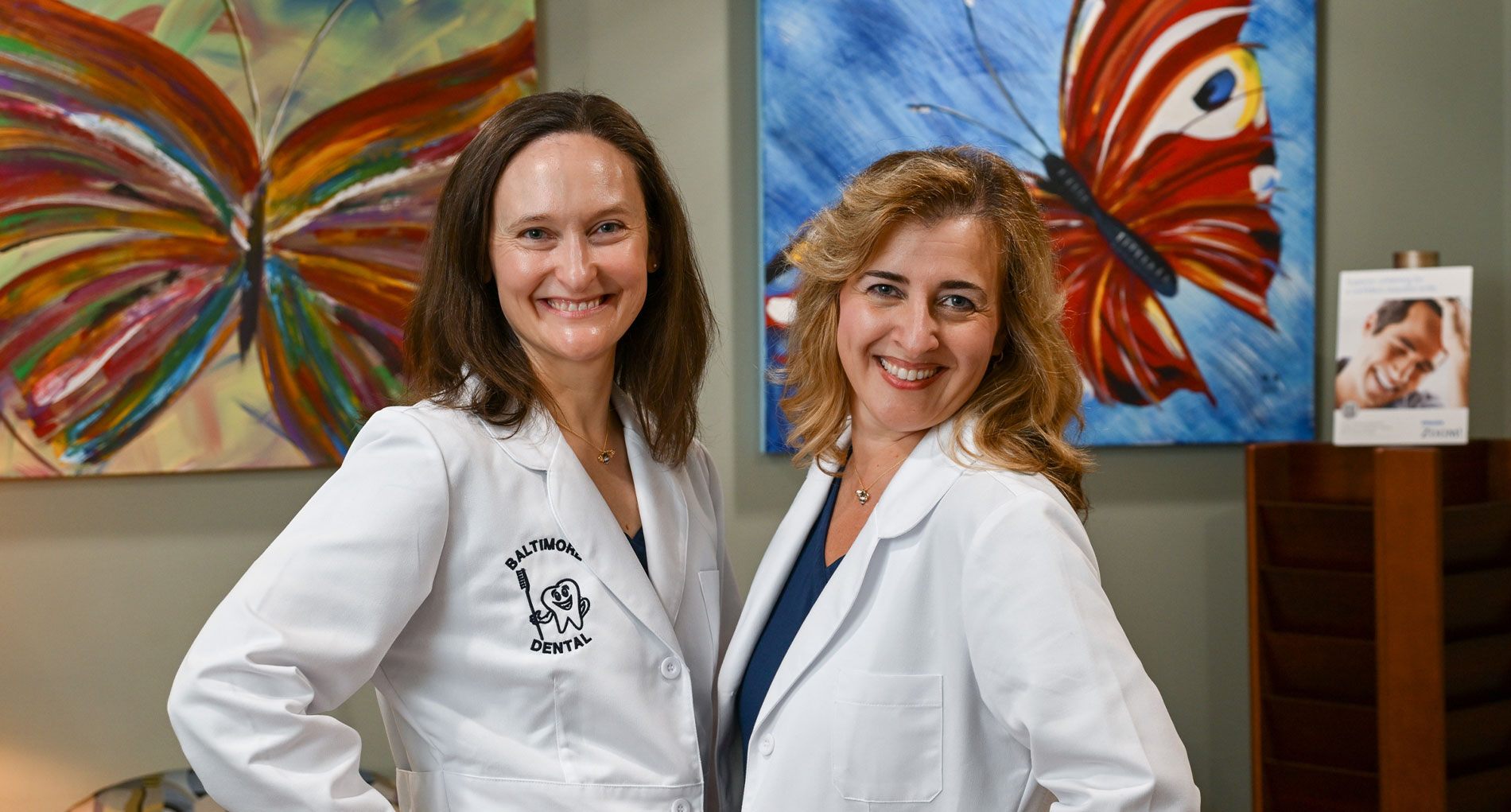 The image shows two individuals, likely professionals, standing in front of a wall with artwork. They are dressed in white lab coats, suggesting they may be scientists or medical professionals.