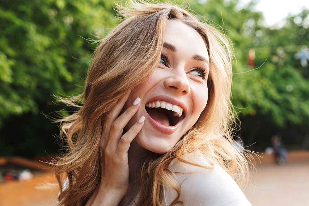 A joyful young woman with a radiant smile, wearing a light-colored top and sunglasses, stands outdoors against a blurred background.