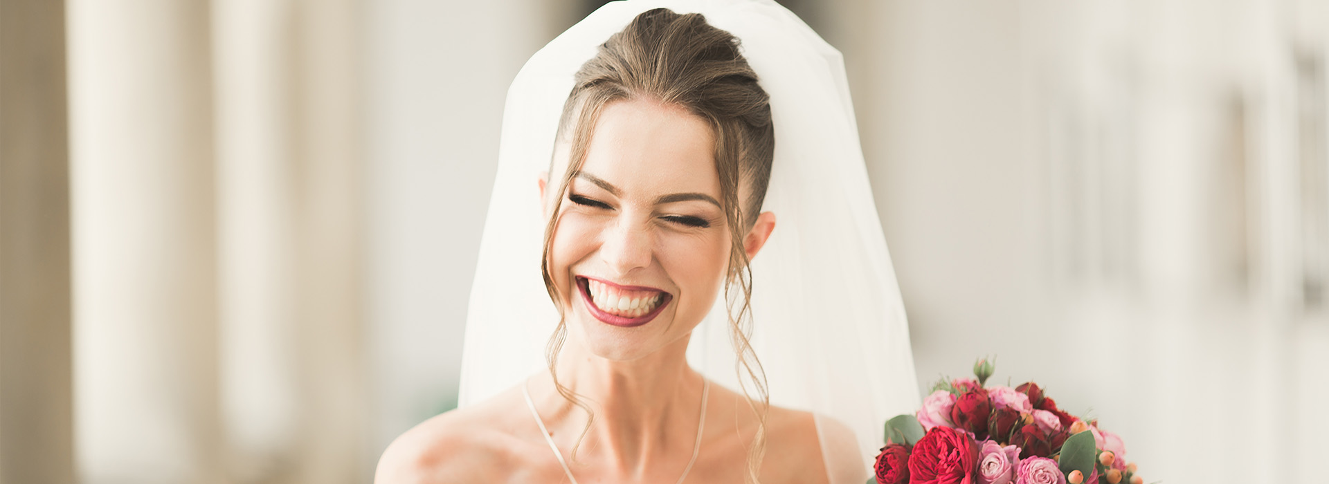 The image shows a woman wearing a wedding veil and smiling, with her eyes closed, while standing in front of a blurred background that suggests an indoor setting.