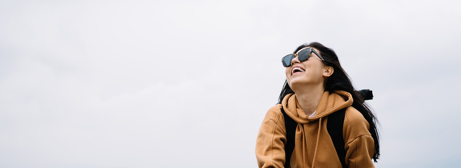 The image shows a person standing outdoors, smiling and looking off to the side, with sunglasses on their head, wearing a backpack, and dressed in casual clothing.