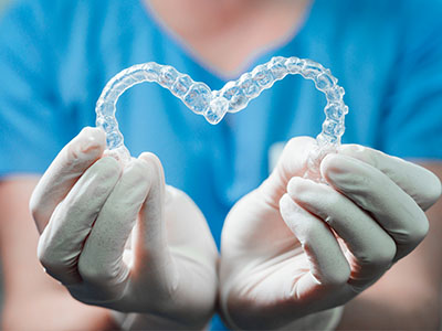 The image shows a person wearing medical gloves and holding a clear plastic heart-shaped object, which appears to be a dental impression or model.