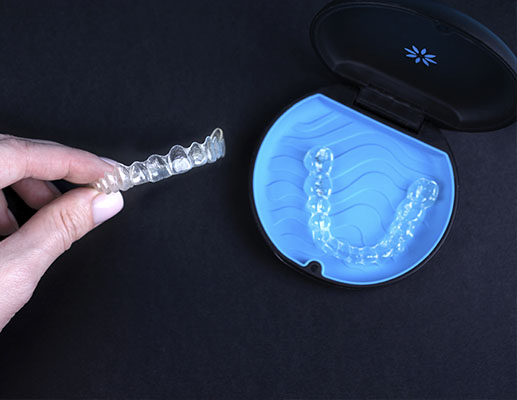 An image of a hand holding a clear dental retainer with a blue container in the background, showcasing the product.