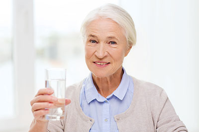 The image features an elderly woman in a domestic setting, holding up a clear glass of water to her mouth with a smile on her face.