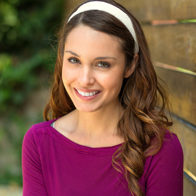 The image is a portrait of a smiling woman with long hair, wearing a purple top and headband, posing against a wooden fence.