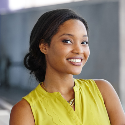 A young woman with a bright smile, wearing a yellow top, stands against a backdrop of a building.