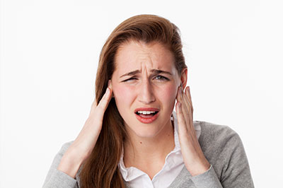 A woman with light brown hair, wearing a white blouse and a gray cardigan, has her hand over her ear and is holding her head in apparent discomfort or pain.