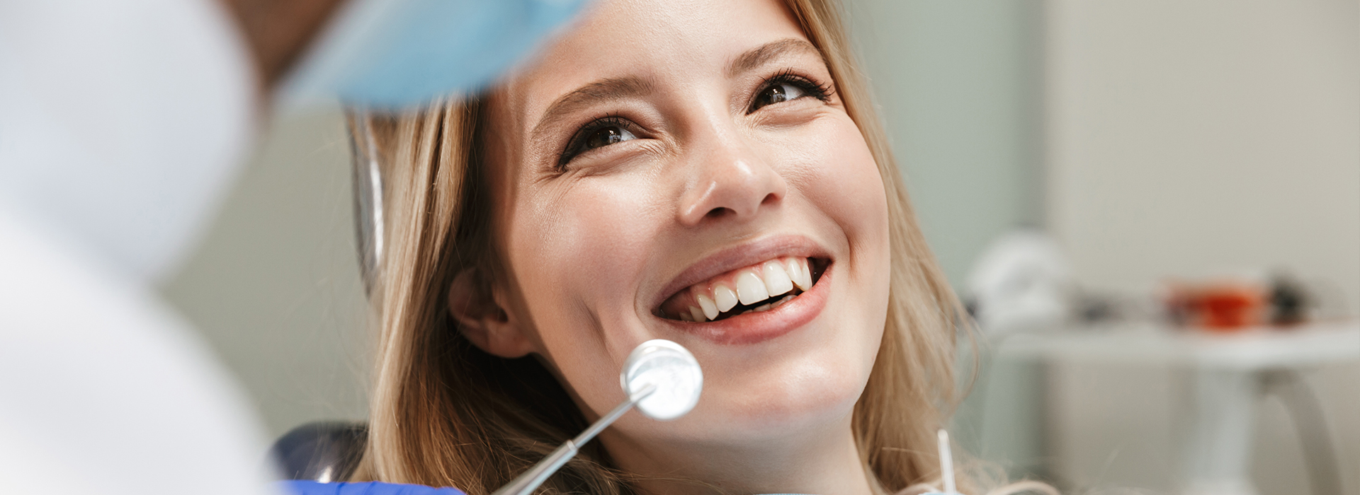 In the image, a woman is smiling at the camera while sitting in a dental chair with a dentist standing nearby. The setting appears to be a dental office, and the woman seems relaxed and comfortable during her visit.