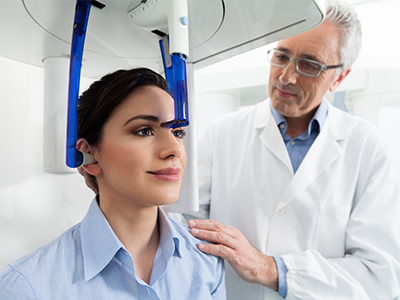 The image shows a woman seated in a dental chair with her head positioned under a device that appears to be used for dental imaging or treatment, while a dentist stands nearby, wearing a lab coat and glasses, observing the equipment.