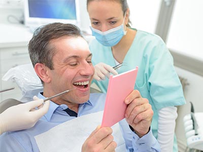 A man in a dental chair, holding a pink card with a surprised expression, while a dentist and hygienist look on.