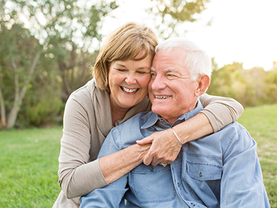 An elderly couple embracing and smiling, with the man in a wheelchair and both wearing casual attire.