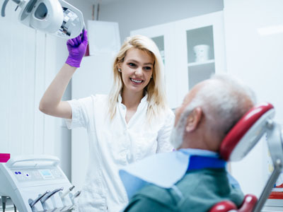 A dental professional is assisting an older man in a dental chair, with the woman holding up a mirror.