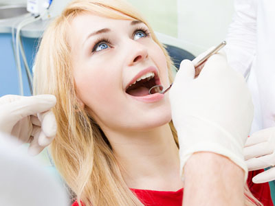 A woman is seated in a dental chair, with her mouth open and a dental professional attending to her. She appears to be receiving dental treatment.