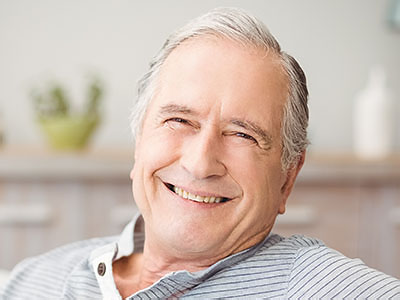 This is a portrait of an elderly man with gray hair, smiling at the camera. He appears to be indoors and is wearing a blue shirt.