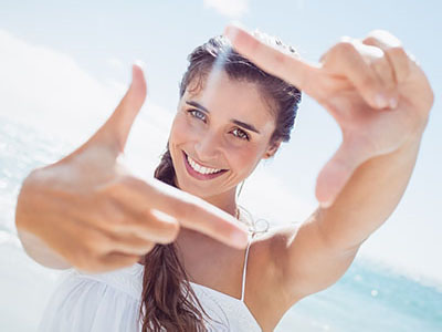 A smiling woman taking a selfie with her hand, against a clear blue sky background.